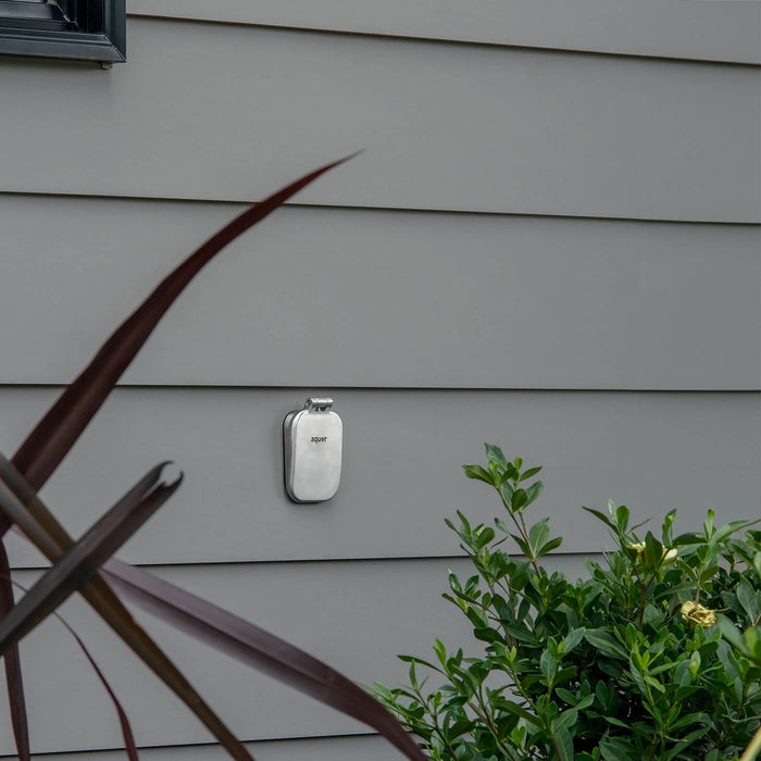 A brushed stainless v2+ debris cover shown on gray lap siding.|47730985795834